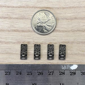 May include: Four small, rectangular, dark metallic buttons featuring an intricate embossed floral pattern and a single central hole. A Canadian 25-cent coin and a metal ruler are included for scale, showing the buttons measure approximately 0.5 inches.