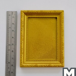 May include: A gold-coloured picture frame with an ornate design. The frame is approximately 19 cm tall and 13 cm wide.