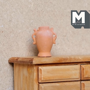 May include: A small terracotta vase with two handles sits on a wooden dresser with three drawers. The vase is a light brown colour and has a slightly textured surface.