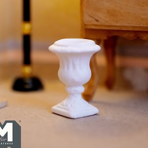 Pode incluir: Urna de cerâmica branca com um design canelado e uma base quadrada. A urna é uma réplica em miniatura de uma urna tradicional.