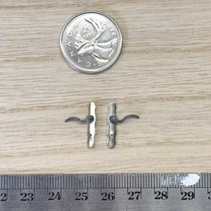 May include: Two miniature silver-toned metal door handles with curved levers and rectangular backplates, positioned above a metal ruler showing a length of approximately 0.5 inches for each handle, next to a US quarter coin for scale.