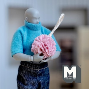 Pode incluir: Uma boneca vestindo um suéter azul de gola alta e jeans azuis está segurando um pompom rosa com uma corda branca presa. A boneca está em pé em frente a uma parede branca com um fundo cinza. A imagem inclui o texto "M for Miniatures".