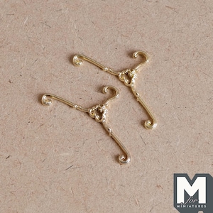 May include: Two gold-colored metal hangers with ornate details. The hangers are designed for miniature dollhouse decor.