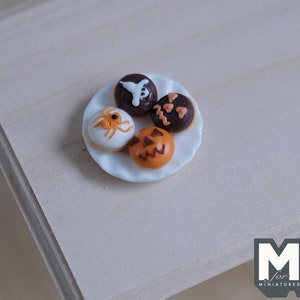 May include: A white plate with four Halloween-themed mini donuts. The donuts are decorated with a ghost, a spider, a pumpkin, and a jack-o'-lantern.