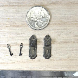 May include: Two miniature dark metal skeleton keys and two matching ornate door plates with knobs. A 2016 Canadian 25-cent coin is positioned above the items for scale, and a metal ruler is placed below, showing the items are under 1 inch long.