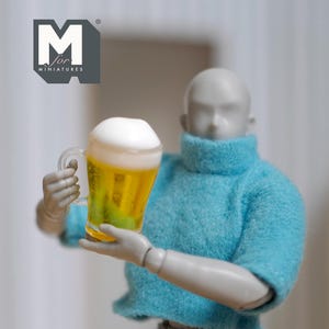 Könnte beinhalten: Ein Miniatur-Bierkrug gefüllt mit goldener Flüssigkeit und bedeckt mit weißem Schaum. Der Krug wird von einer grauen, beweglichen Figur gehalten, die einen blauen Pullover trägt. Das Logo "M for MINIATURES" befindet sich in der oberen linken Ecke.