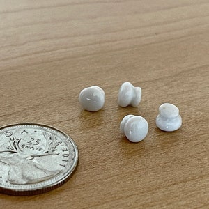 Peut inclure: Quatre boutons en céramique blanche avec une surface lisse et arrondie. Les boutons sont disposés en demi-cercle sur une surface en bois. Une pièce de monnaie canadienne de 25 cents se trouve dans le coin inférieur gauche de l'image.