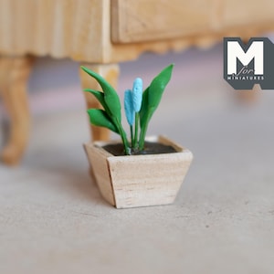 May include: A miniature wooden planter with a light brown finish, filled with green leaves and blue flowers. The planter is labelled "M for Miniatures".