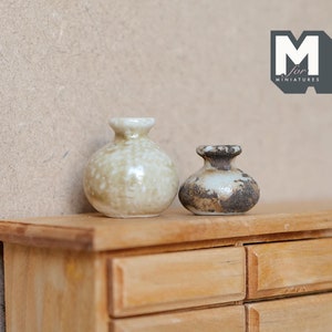 May include: Two small ceramic vases, one with a white glaze and brown speckles, the other with a brown glaze and white speckles, sitting on a wooden dresser with drawers.