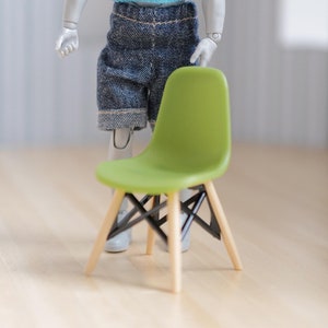 Può includere: Una bambola in miniatura che indossa un maglione blu chiaro e pantaloncini di jeans blu si trova accanto a una sedia verde con gambe in legno. La sedia ha un design semplice e moderno.