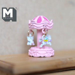 May include: A miniature pink carousel with a striped roof and two hanging unicorn figures. The carousel has a decorative base and is designed for miniatures. The carousel is made of a light pink material with a darker pink roof.
