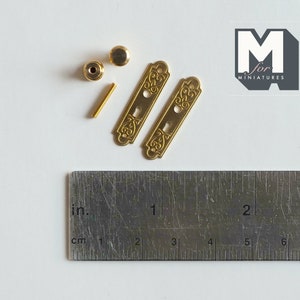 May include: Two gold-toned door hardware plates with ornate designs, two gold-toned knobs, and a gold-toned screw. The hardware is measured against a ruler with inches and centimeters.