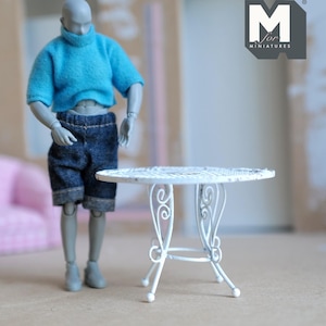 May include: A miniature white metal table with a round top and decorative scrollwork on the legs. The table is next to a doll wearing a blue sweater and denim shorts. The logo "M for MINATURES" is in the upper right corner.