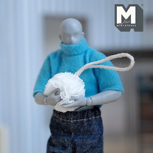 Könnte beinhalten: Eine graue Actionfigur trägt einen blauen Rollkragenpullover und eine blaue Jeans und hält einen weißen Pompon mit einer daran befestigten Schnur. Die Figur steht vor einer weißen Wand mit einem Logo, das "M for Miniatures" lautet.