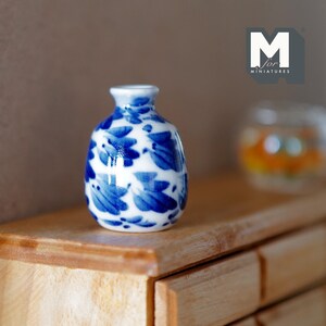 Könnte beinhalten: Eine kleine, weiße Keramikvase mit einem blauen und weißen Blumenmuster. Die Vase steht auf einer Holzoberfläche.