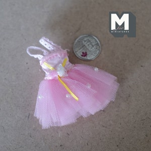 Puede incluir: Un vestido de tul rosa con un corpiño de satén blanco y una cinta amarilla. El vestido tiene un adorno de concha y detalles de perlas.