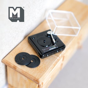 May include: A miniature black record player with a clear dust cover and two black vinyl records. The record player has a silver tone arm and a small control panel with a rainbow equalizer graphic. The text "M for Miniatures" is visible in the top left corner of the image.