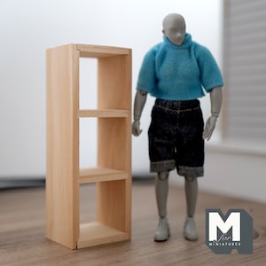 May include: A small, unfinished wooden bookshelf with three shelves stands next to a miniature mannequin wearing a blue hoodie and denim shorts. The bookshelf is made of light-colored wood and is designed for miniature items.