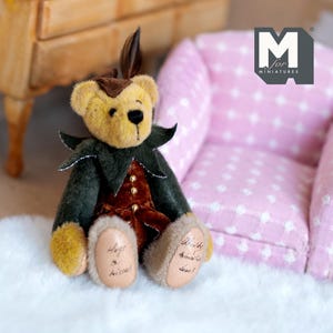 May include: A small, plush teddy bear wearing a green and brown jester costume. The bear is sitting on a white, fluffy surface. The bear's paws have the words "Angel" and "Whisker" written on them.