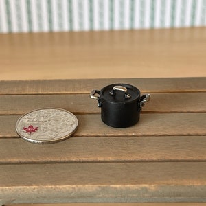 May include: A miniature black pot with a silver lid and handles. The pot is sitting on a wooden surface next to a Canadian coin.