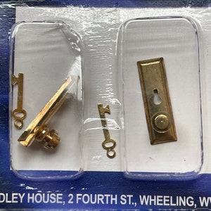 以下が含まれることがあります： 2つの鍵、ドアハンドル、ドアノブがついたドールハウス金具キットのクローズアップ。キットは、青と白の背景の透明なプラスチック製のブリスターパックに梱包されています。パッケージには「By Handley」と「QUALITY DOLLHOUSE SUPPLIES」の文字が印刷されています。パッケージの下部には「DLEY HOUSE, 2 FOURTH ST., WHEELING, W」の文字が印刷されています。