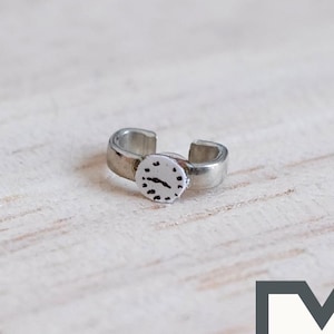 Op de afbeelding: Een zilverkleurige miniatuurhorloge-ring met een wit gezicht en zwarte markeringen. De ring is aan één kant open en kan aan een vinger worden gedragen.