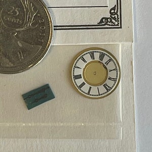 May include: A small, white clock face with black Roman numerals and a gold rim. The clock face is surrounded by a white background.