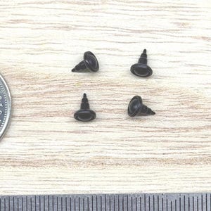 May include: Four small black plastic safety eyes for crafts, each featuring a rounded head and a pointed post. A Canadian 25-cent coin is positioned to the left for scale, and a metal ruler is placed at the bottom to show the size in inches.