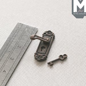 May include: A miniature bronze-toned metal door handle plate with an ornate floral design and a matching small key. A metal ruler is positioned alongside the items, showing the handle plate measures approximately 3.2 cm in length.