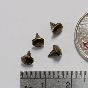 May include: Four small gold-coloured metal screws with hexagonal heads, measuring approximately 1.5 cm in length, are arranged on a white surface next to a ruler and a Canadian 25-cent coin.