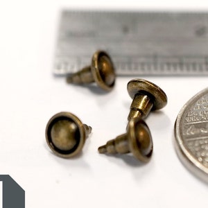 May include: Three antique gold-toned metal dome-shaped tacks with a pointed end, next to a Canadian 25-cent coin and a ruler.