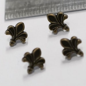 May include: Four antique gold-toned metal fleur-de-lis embellishments. The embellishments are approximately 2.5 cm in size.
