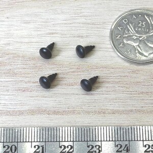 May include: Four small black metal screws with rounded heads arranged in a square formation. A silver Canadian 25-cent coin is positioned to the right, and a metal ruler showing measurements in inches and centimeters is placed along the bottom edge.