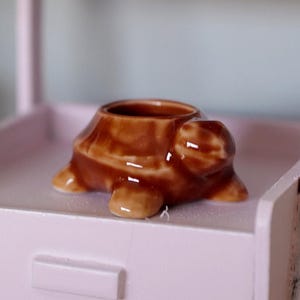 Pode incluir: Um pequeno vaso em forma de tartaruga de cerâmica marrom. A tartaruga tem as costas escavadas para plantar pequenas suculentas ou ervas. O vaso está sobre uma superfície rosa claro.