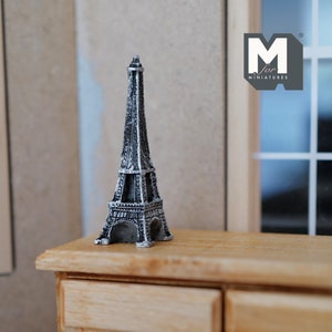 Puede incluir: Una réplica en miniatura de la Torre Eiffel hecha de metal con un acabado de pintura en blanco y negro. La torre está sentada sobre una superficie de madera.