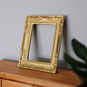 May include: A gold-colored ornate picture frame with a decorative border. The frame is sitting on a wooden surface.