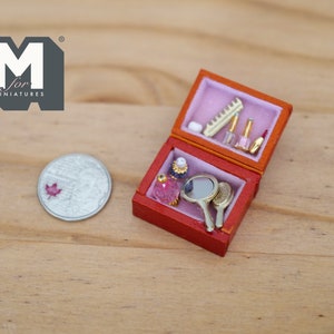 Puede incluir: Un estuche de tocador en miniatura de color rojo y dorado con un interior rosa. El estuche está abierto y contiene un peine, un frasco de perfume, un lápiz labial y un pequeño espejo.