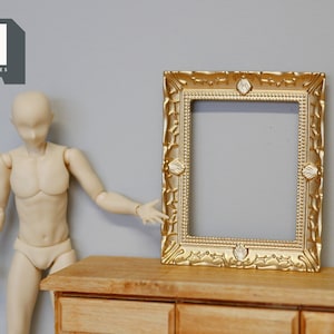 May include: A gold-colored ornate picture frame with a detailed design. The frame is sitting on a wooden table.