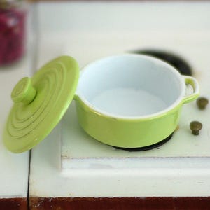 Pode incluir: Uma panela de cerâmica verde limão em miniatura com sua tampa correspondente, colocada em uma superfície branca. A panela tem um interior branco e pequenas alças. A tampa está de lado, revelando o interior da panela.