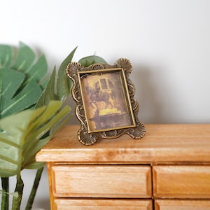 May include: A small, ornate, gold-toned picture frame with a decorative design. The frame is sitting on a wooden surface with drawers.