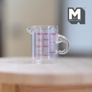 May include: A clear plastic miniature measuring cup with a handle and a pouring spout. The cup has pink measurement markings for cups and ounces. The logo "M for Miniatures" is in the upper right corner.