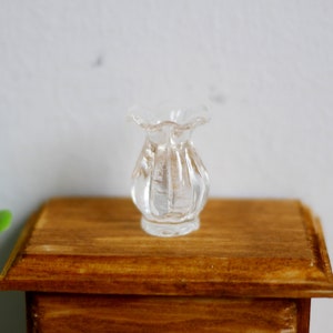 Op de afbeelding: Een kleine, heldere glazen vaas met een gerimpelde rand. De vaas heeft verticale groeven en staat op een houten oppervlak. Deze miniatuurvaas is waarschijnlijk voor een poppenhuis of een vergelijkbare miniatuurdisplay.