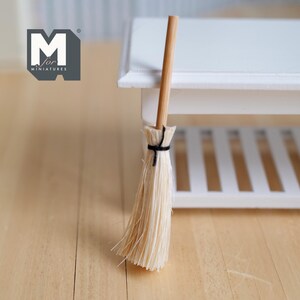 Può includere: Una scopa in miniatura con un manico in legno marrone chiaro e setole di paglia. Le setole sono legate con spago nero. La scopa è posizionata accanto a un tavolino bianco in miniatura. Il logo "M for miniatures" è nell'angolo in alto a sinistra.