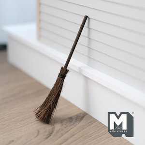 Può includere: Una scopa marrone in miniatura con un manico di legno, appoggiata a un muro bianco. La scopa è lunga circa 25 cm.
