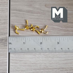 May include: Four gold-colored metal hooks, each with a single hook at the end, are arranged on a wooden surface. A ruler is placed below the hooks, showing that they are approximately 1 inch in length.