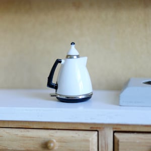 May include: A miniature white electric kettle with a black handle and silver accents. The small appliance has a rounded shape and a black and white top. It is set on a white surface.