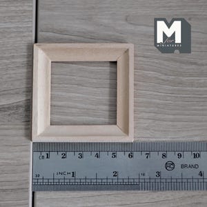 May include: A small, unfinished wooden square frame, ideal for miniature projects. The frame is light-coloured and has a simple design. A ruler with centimetre and inch measurements is placed next to the frame. The text "M for MINIATURES" is in the upper right corner.