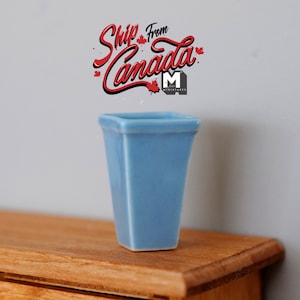 May include: A light blue ceramic vase with a square base and a slightly flared top. The vase is sitting on a wooden surface.