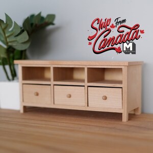 May include: A miniature wooden cabinet with three drawers and open shelves. The cabinet is made of light-colored wood and has a simple, modern design. A potted plant is to the left of the cabinet. Text on the wall says "Ship From Canada".