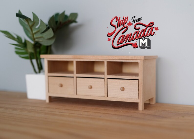 May include: A miniature wooden cabinet with three drawers and open shelves. The cabinet is made of light-colored wood and has a simple, modern design. A potted plant is to the left of the cabinet. Text on the wall says "Ship From Canada".